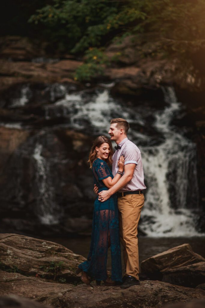 Mom and dad standing close together on broad rocks with a cascading waterfall behind them.