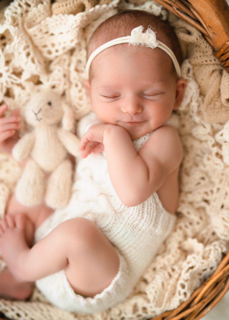 Smiling newborn baby girl holds a cream bunny stuffy wearing a white knit romper and laying in a wooden bowl.