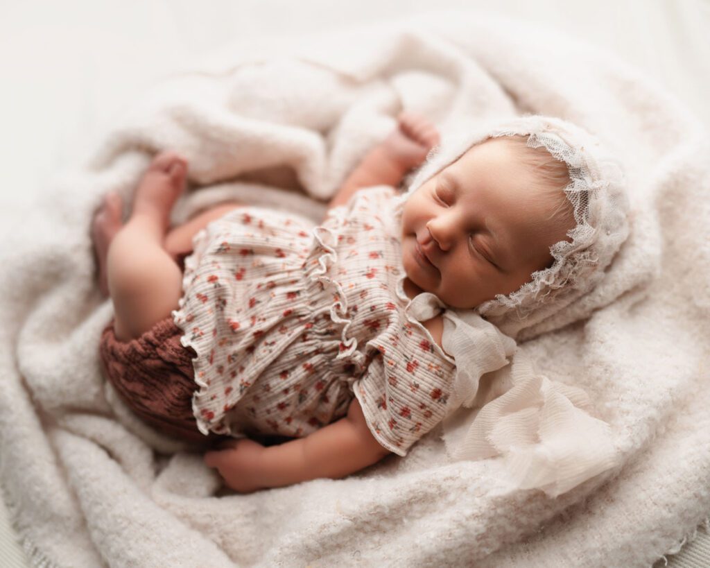 Baby girl sleeping on a soft plush blanket wearing a pink floral shirt and bloomers with a soft smile on her face.