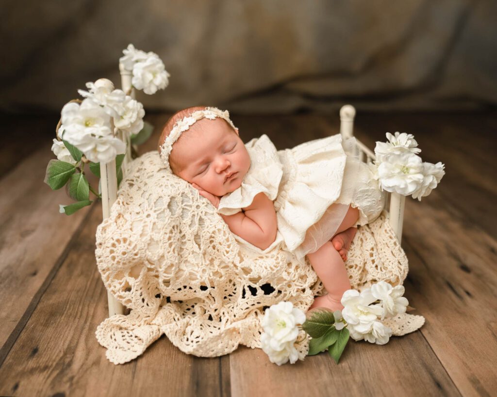 Sleeping baby girl wearing a white dress lays on a vintage crochet blanket on an iron bed covered with flowers.