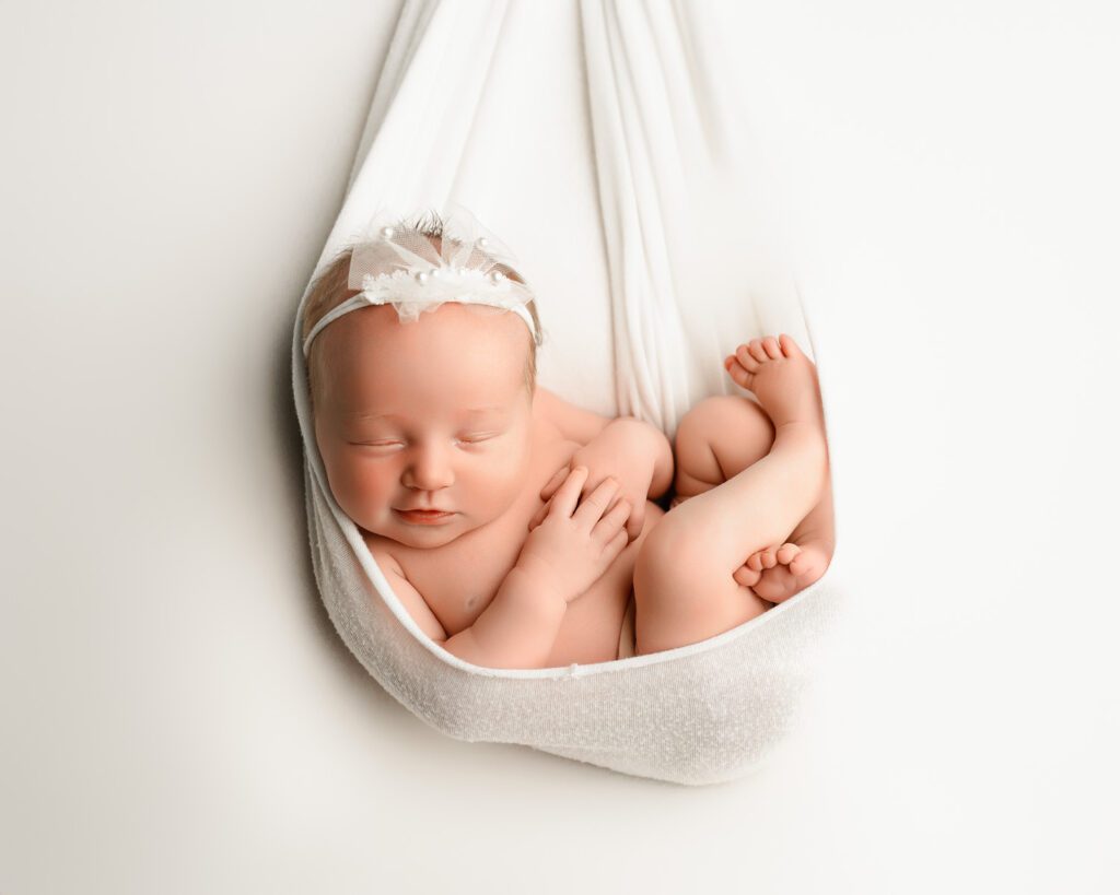 White stork swaddle swing holding a newborn baby girl wearing a white pearl headband curled up in a bundle.