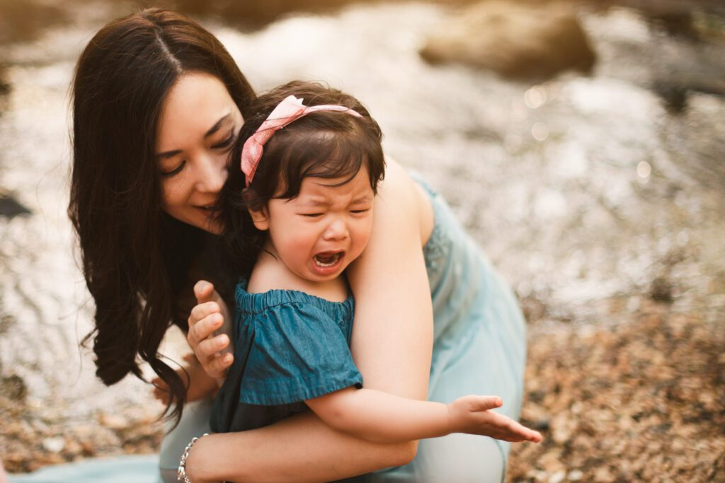 Mom holding and comforting her young daughter beside the creek while the child reaches outward.
