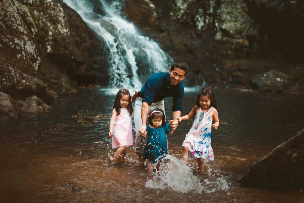 Dad walking through shallow creek water with his three daughters as they splash around him.