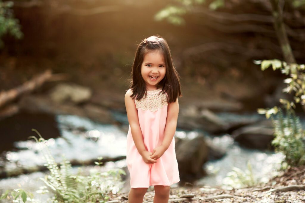Young girl in a pink dress standing beside the creek with smooth rocks and flowing water behind her.