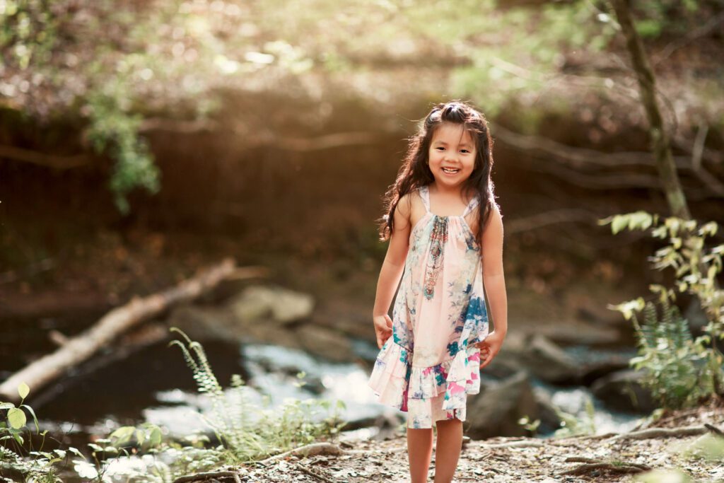 Smiling daughter in a floral dress standing near the creek with rocks and woodland greenery behind her.