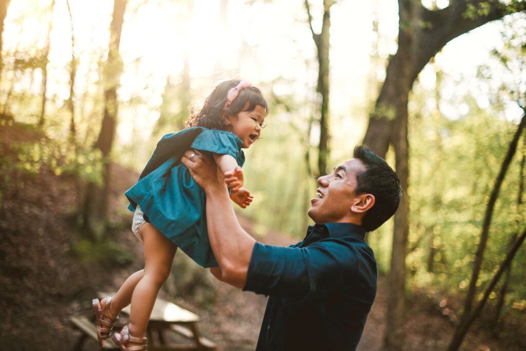 Dad lifting his young daughter into the air beneath tall trees with warm sunlight filtering through the woods.