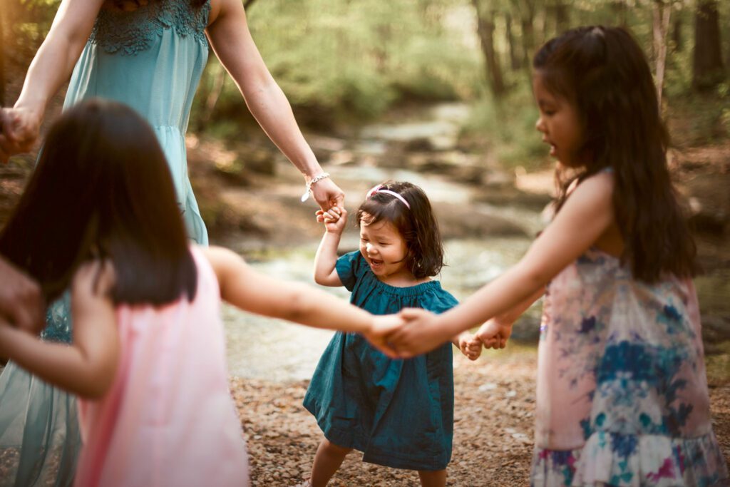 Three sisters holding hands in a circle beside the shallow creek with a wooded bank in the background.