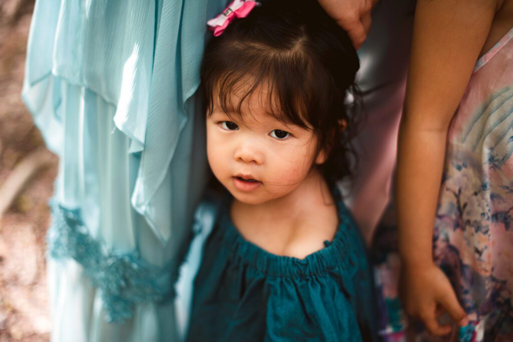 Young daughter in a teal dress standing close between mom and her sisters in soft woodland light.