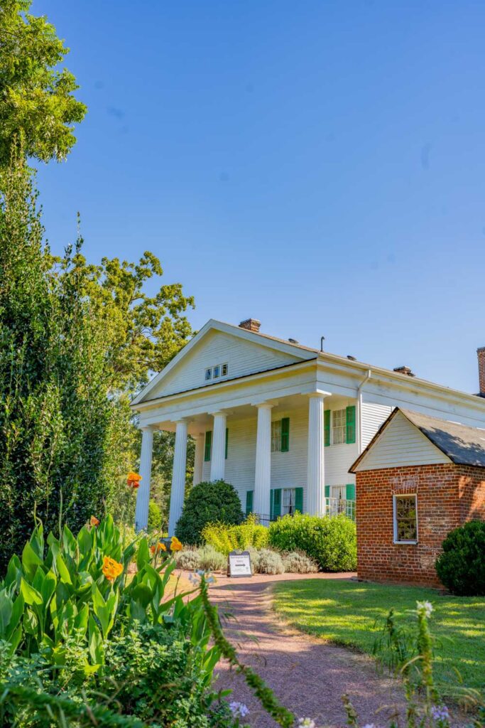 Garden path and orange flowers beside Barrington Hall’s white-columned exterior and a small brick building
