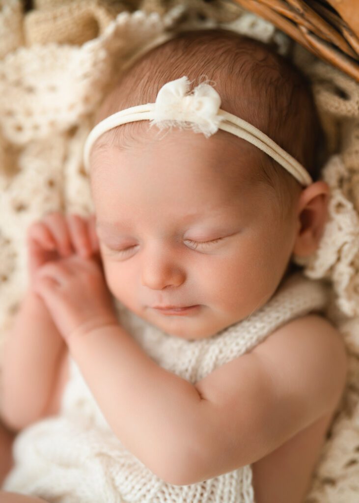 Sleeping newborn baby girl in a knit onesie and bow headband with both hands resting near her face.