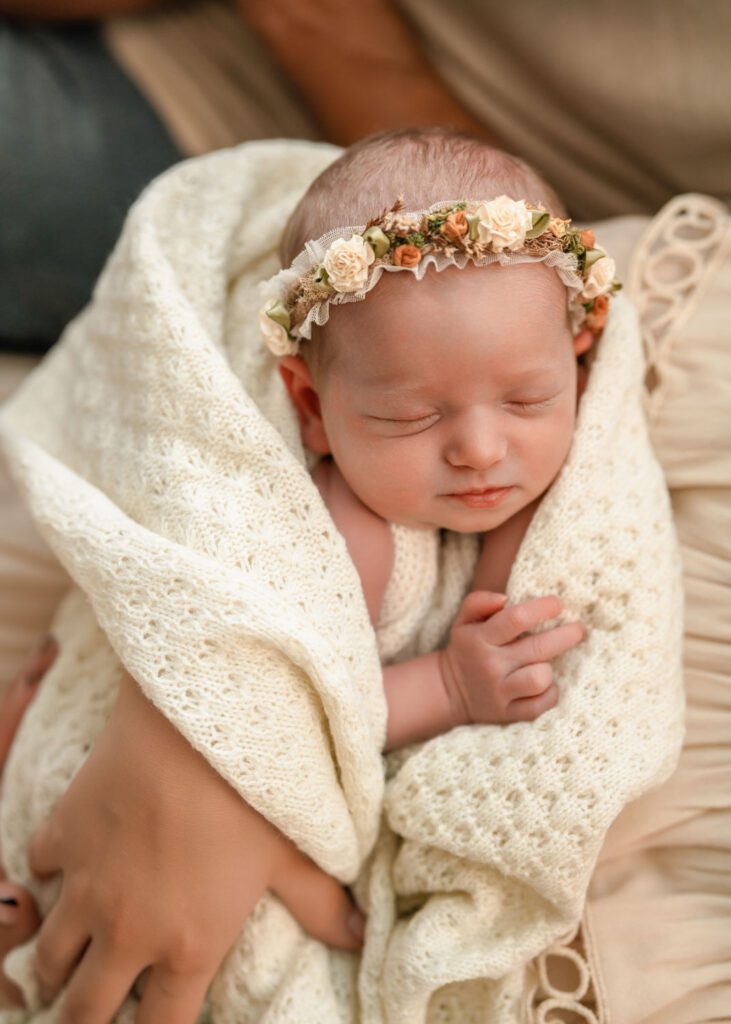 Newborn baby girl in a floral headband wrapped in a cream knit blanket while resting in her mother’s arms.