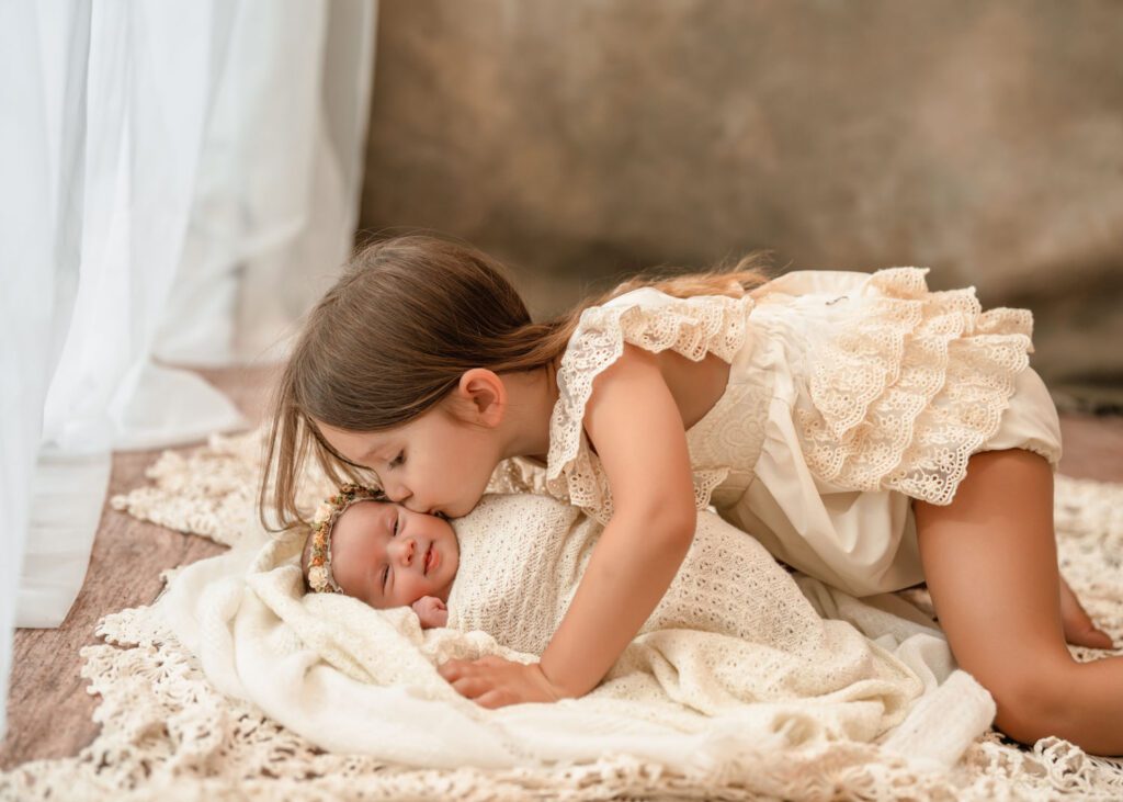 Big sister kissing her newborn baby sister on the forehead as the baby rests wrapped in cream blankets.