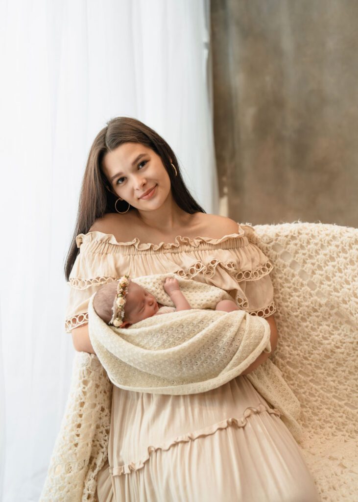 Mom holding her newborn daughter beside a bright window as baby wears a neutral floral headband.