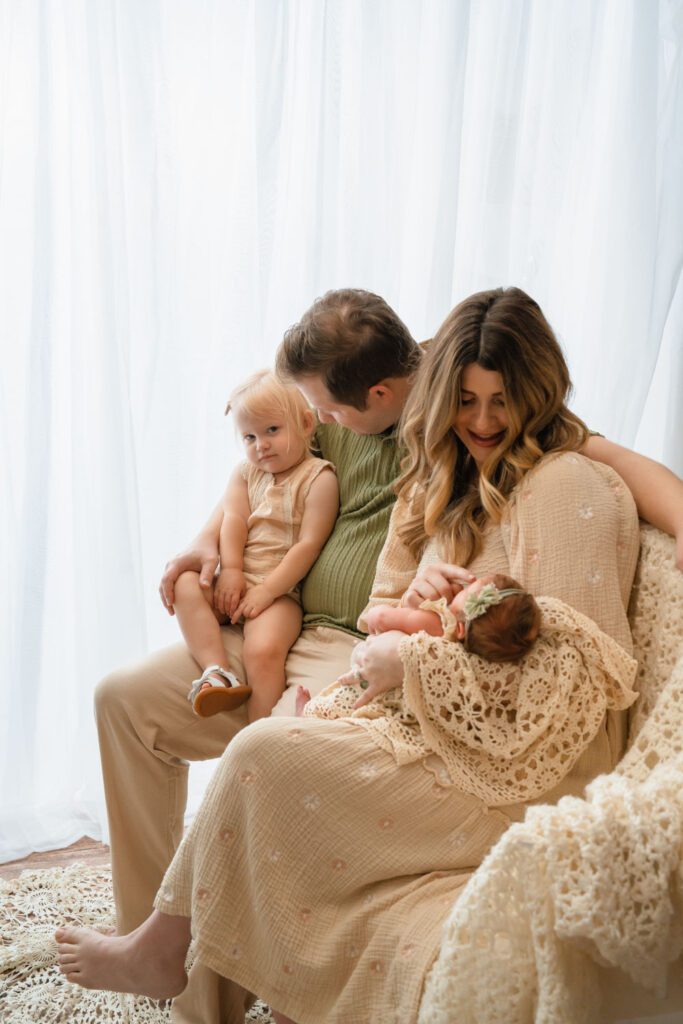 Family sitting beside a bright window as mom holds the newborn and dad holds their toddler daughter.