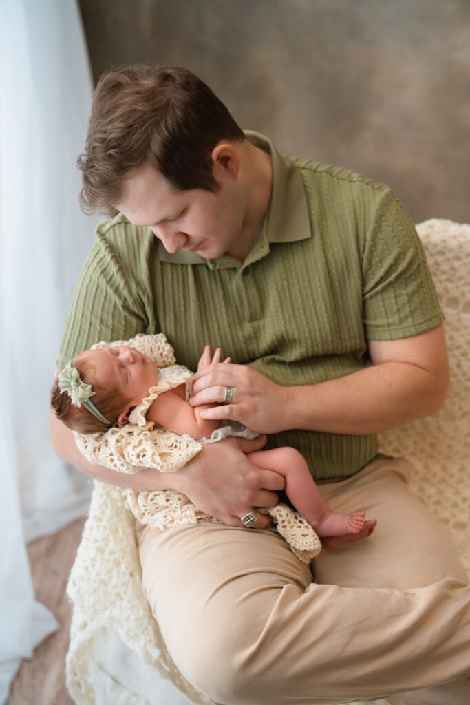 Father in a green shirt holding his newborn daughter in a cream lace outfit and green bow headband.