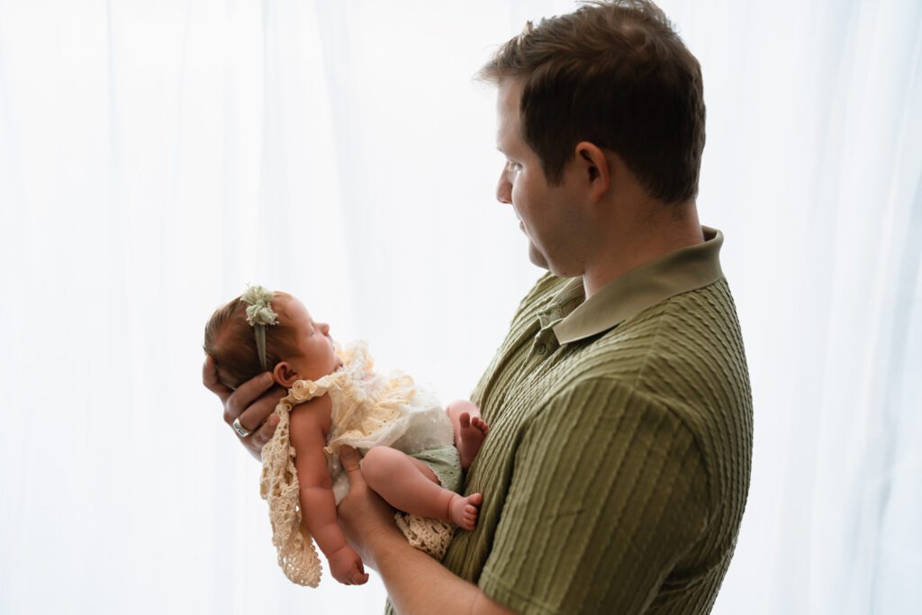 Dad in a green collared shirt holding his newborn daughter wrapped in crochet against a bright white window.