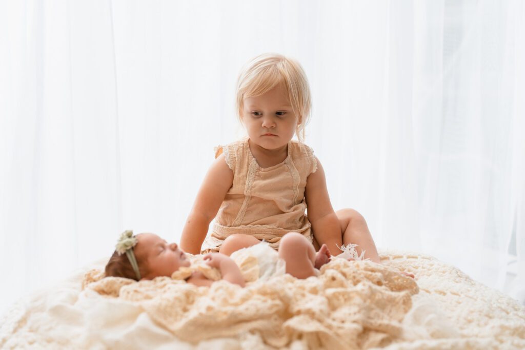 Big sister sitting beside her sleeping newborn sister on layered cream blankets with an uncertain expression.
