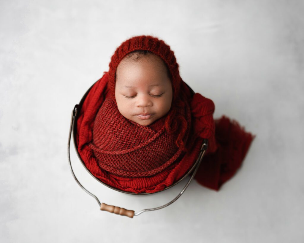 six week old Newborn baby boy swaddled in a red knit wrap wearing a matching bonnet placed in bucket
