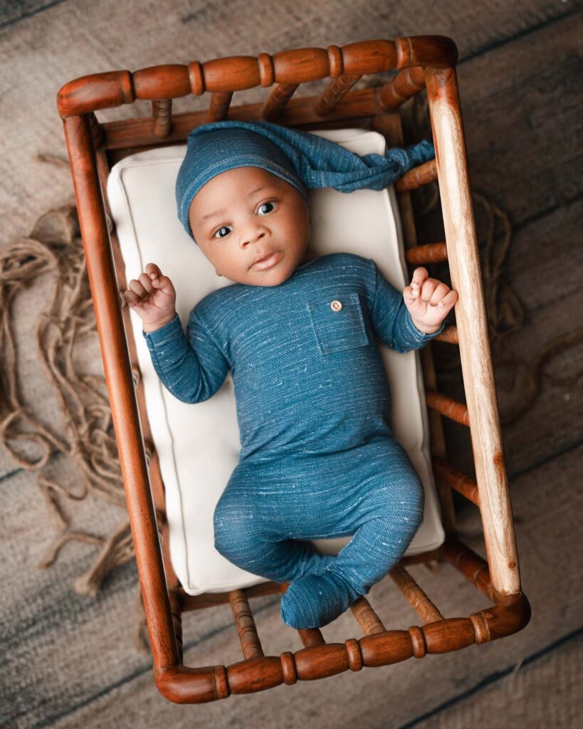 Awake baby boy laying on his back in a wooden spindle crib wearing blue pajamas with a matching sleepy cap hat.
