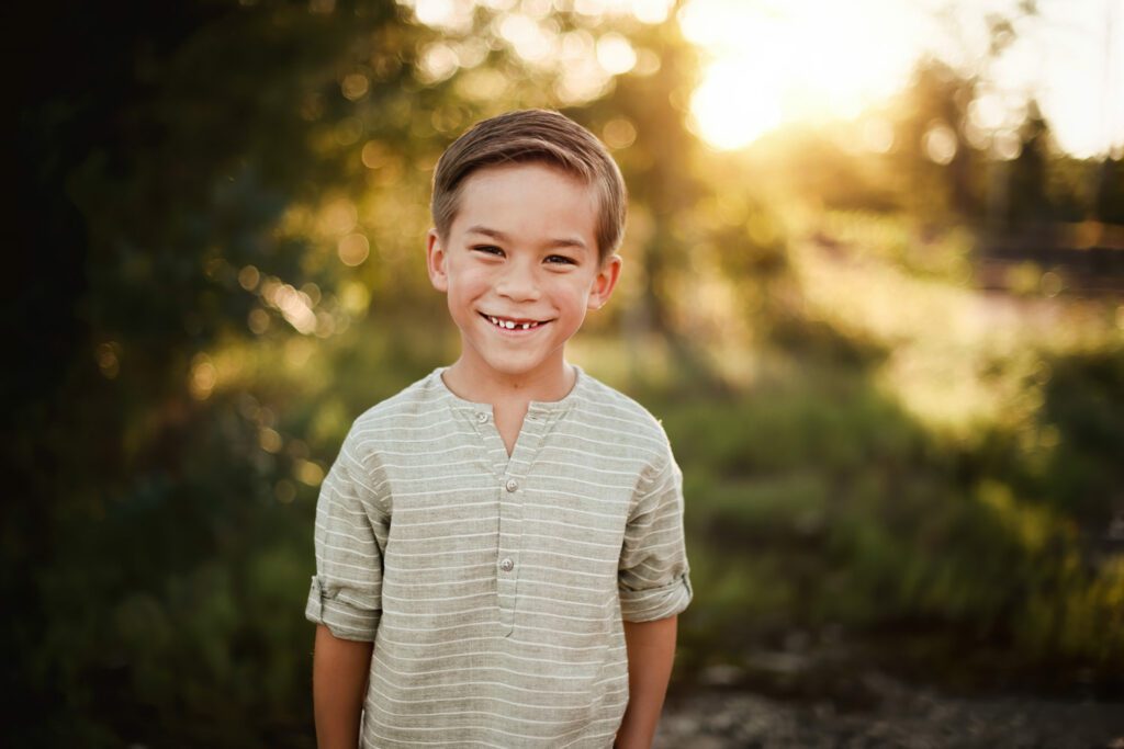 Smiling boy standing in forest against a softly blurred green background in warm golden evening light.