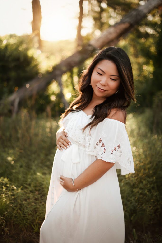Pregnant woman in a white off-the-shoulder gown cradling her belly outdoors among softly lit trees and greenery.