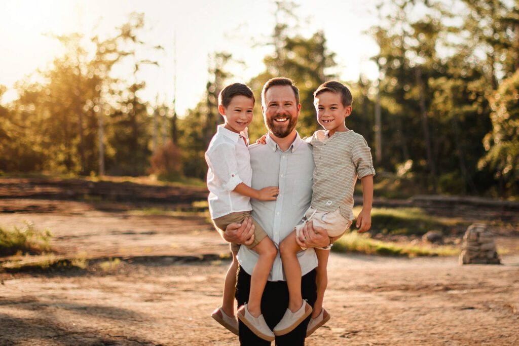 smiling dad holding two twin brothers outdoors in nature and trees near a riverbed.