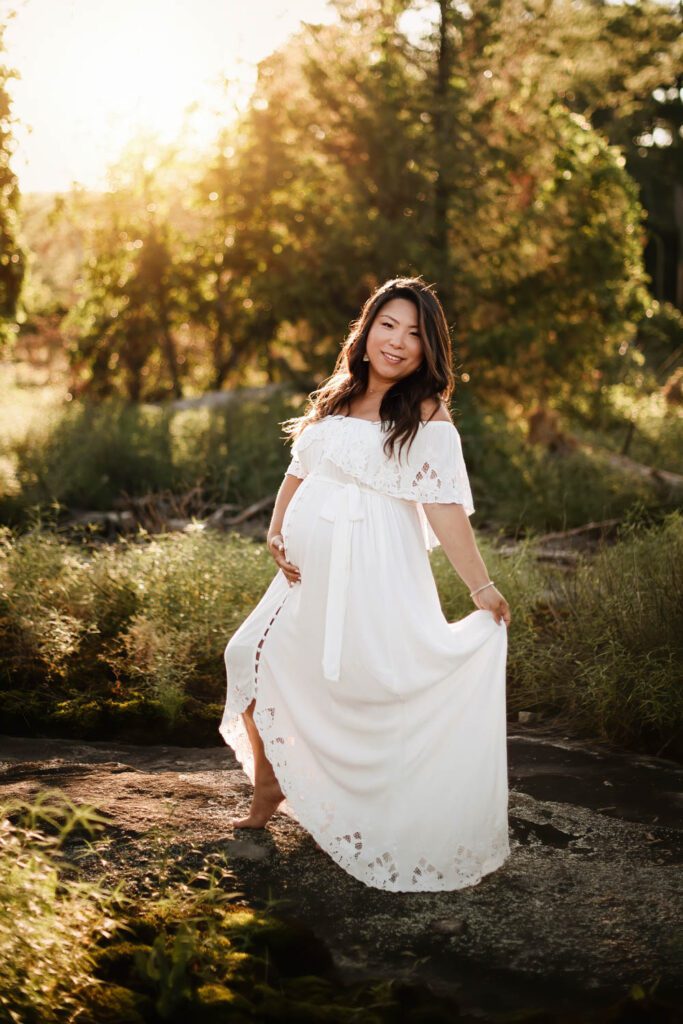 Pregnant woman in a flowing white gown standing alone among golden trees and greenery during an outdoor maternity session.