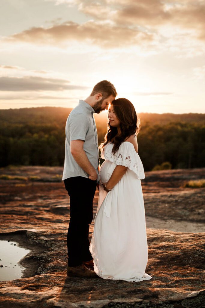 Pregnant woman in a white gown standing close to husband with their foreheads touching in warm sunset light.