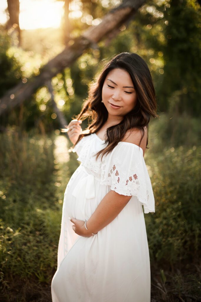 pregnant woman in white eyelet dress standing outdoors in greenery twirling her har and touching her belly.