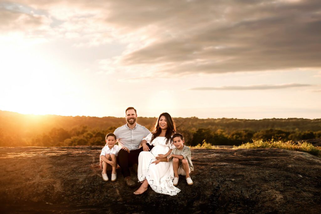 Pregnant woman in a white gown seated with husband and two children on the rocks beneath a glowing sunset sky.