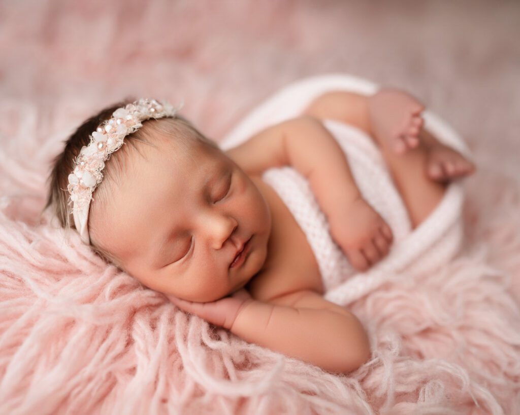 Newborn baby girl laying on a pink fur backdrop swaddled in a pink wrap with a beaded crystal headband in her hair.