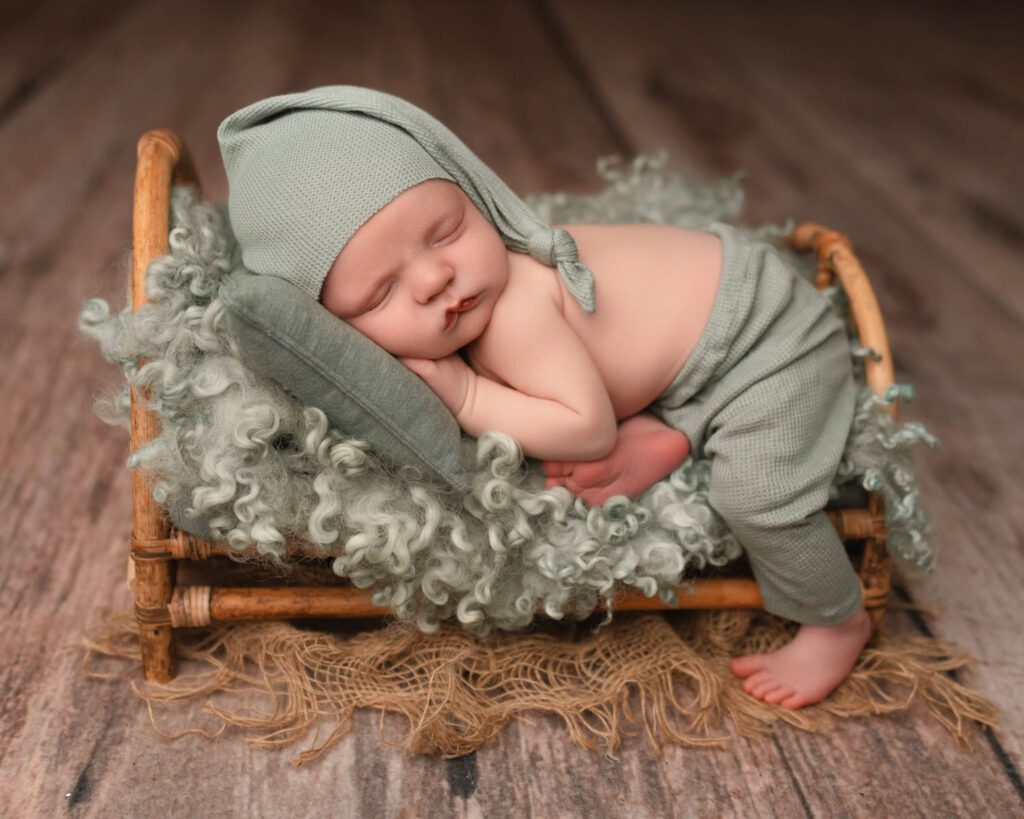 two week old Baby boy wears sage green pants and matching sleepy cap while laying on a bamboo newborn bed.