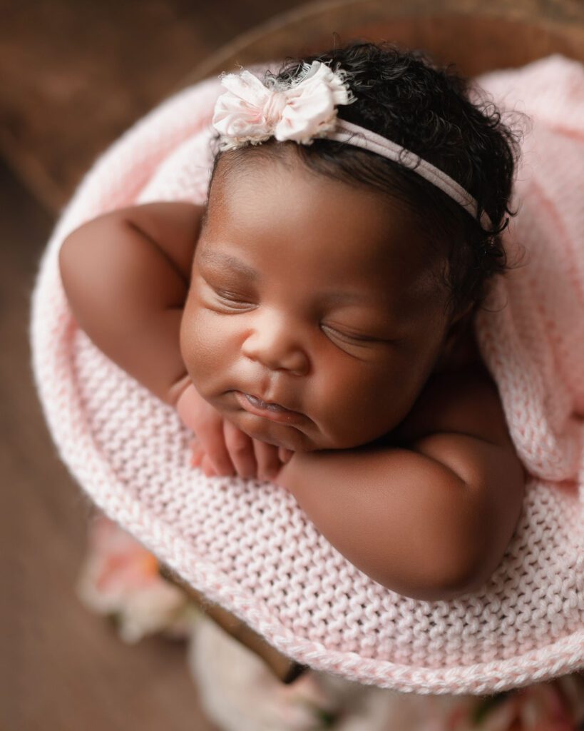 Newborn baby girl wears a pink bow headband in her thick black hair and rests her head on her hands on a pink blanket.
