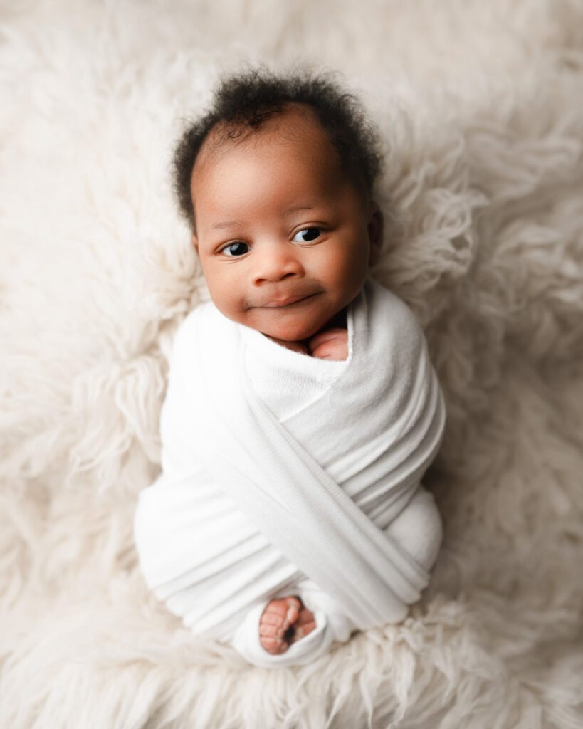 Smiling newborn baby boy with black hair is swaddled in a white wrap laying on a white fur backdrop looks at the camera.