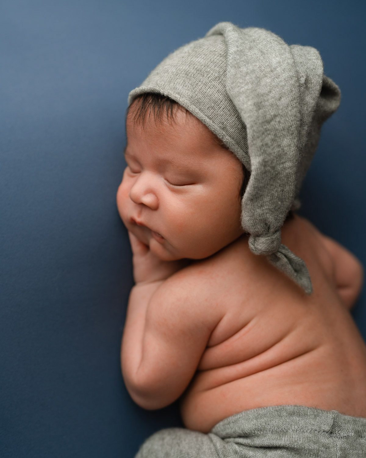 Sleeping newborn baby boy lays on his stomach on a blue backdrop and wears grey pants and a matching sleepy cap hat.