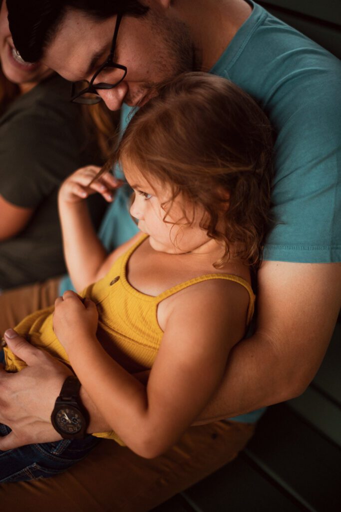 Daughter wearing yellow tank top and blue jeans is sad and unhappy while her dad wearing a blue shirt comforts her.