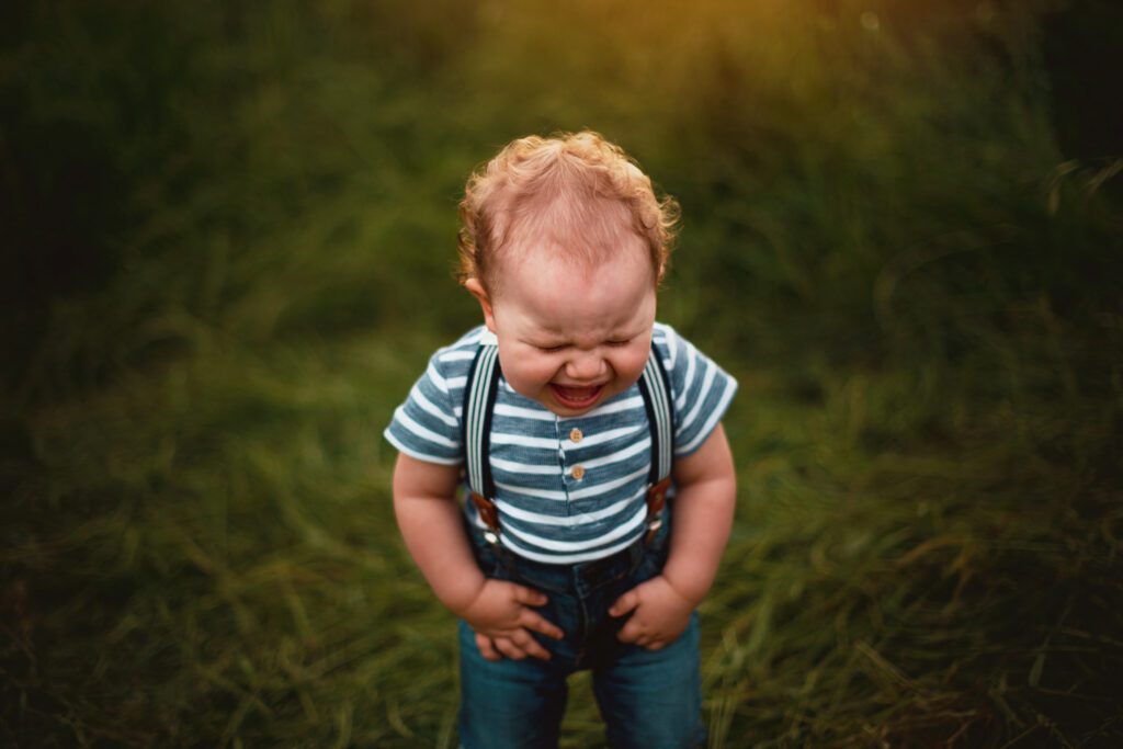 Baby boy in a blue and white striped shirt with suspenders and jeans cries and pitches at fit standing in grass at sunset.