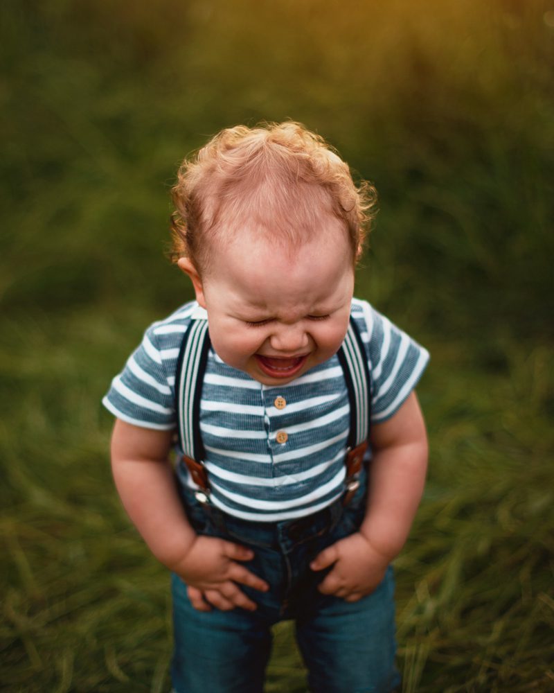 Baby boy in a blue and white striped shirt with suspenders and jeans cries and pitches at fit standing in grass at sunset.