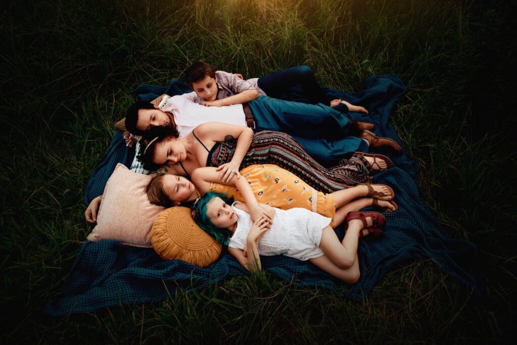 Family laying on a blue blanket in the grass all wearing coordinating outfits in mustard yellow, blue, pink, and browns. 